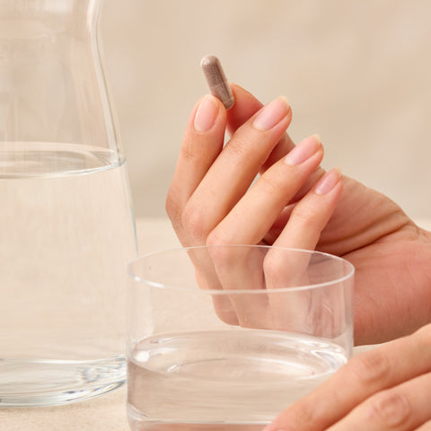 Hand holding a Juice Plus+ capsule next to a glass of water on a neutral background