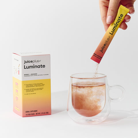 Juice Plus+ Luminate supplement stick pack being added to a glass of water with the product box in the background.
