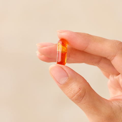 Hand holding a multicolored Juice Plus+ Omega capsule against a beige background
