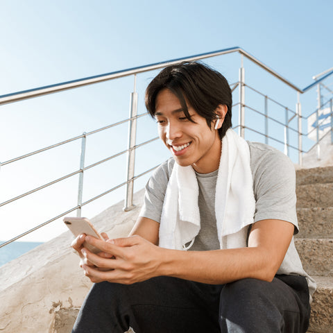 Man sitting outdoors using a phone with a towel around his neck, smiling.