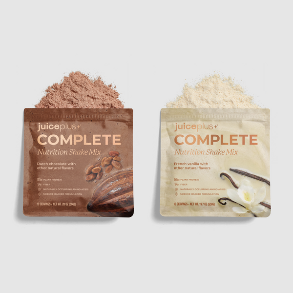 Two Juice Plus+ Complete Nutrition Shake Mix pouches on a light gray background, each with a small mound of powder above it. The left pouch is dark brown chocolate flavor, and the right pouch is light beige vanilla flavor.