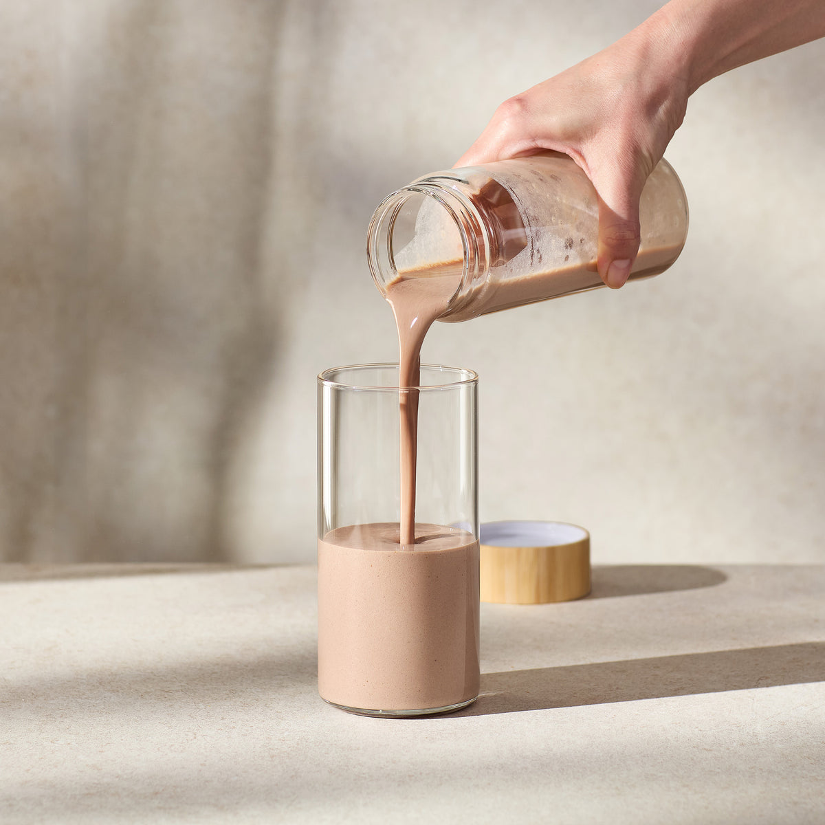 Person pouring a creamy liquid from a glass jar into another glass on a neutral background