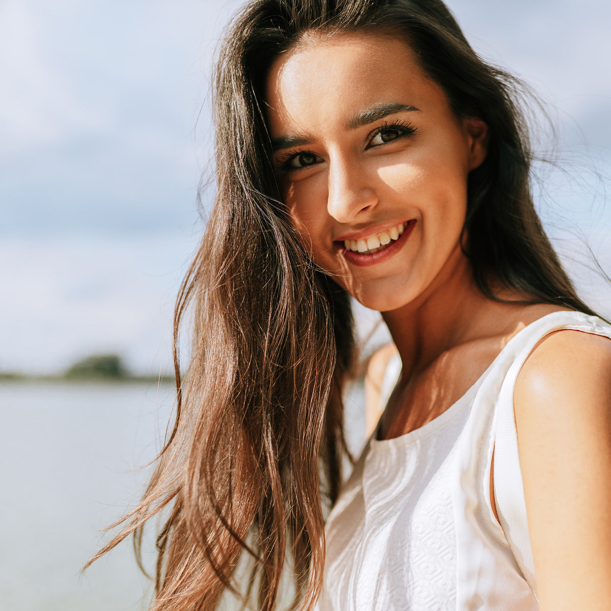 Woman with long brown hair smiling outdoors with a blurred natural background