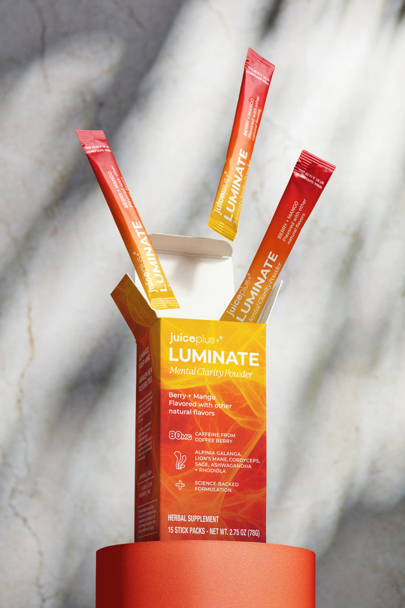 A box of Juiceplus LUMINATE Mental Clarity Powder with three individual stick packs fanned out behind it on a red cylindrical pedestal against a light marble background.