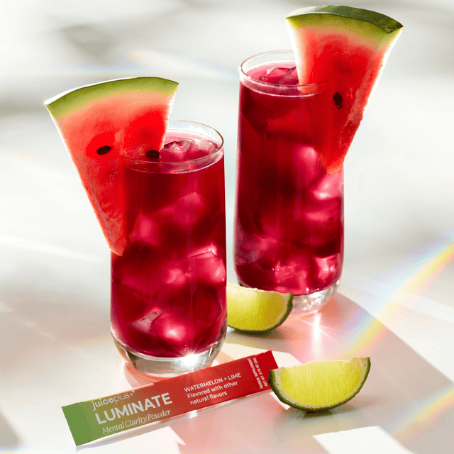 Two tall glasses filled with a bright pink-red iced drink and garnished with watermelon slices, with lime wedges and a juice棒 package in front on a white surface.