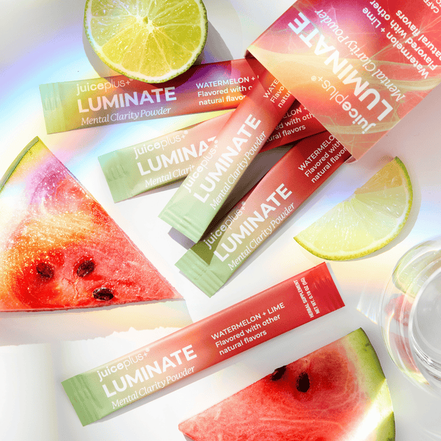 Juiceplus LUMINATE Mental Clarity Powder packets arranged on a white surface with watermelon slices and lime wedges. The packets are red, orange, and green, with text indicating Watermelon + Lime flavor.