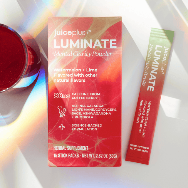 Juiceplus LUMINATE Mental Clarity Powder shown with its red and orange box and a single stick pack on a white surface with a red cup partially visible at left.