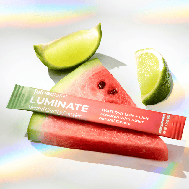 A juiceplus+ Luminate Mental Clarity Powder stick pack in Watermelon + Lime flavor, styled on top of a watermelon slice with lime wedges on a bright white background with rainbow light leaks.