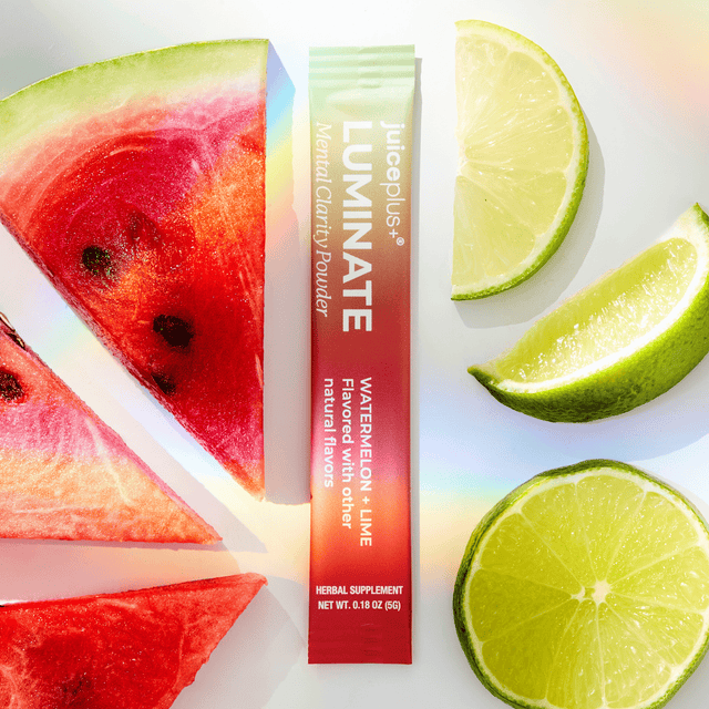 A single packet of JuicePlus+ LUMINATE Watermelon + Lime herbal supplement powder is centered on a white background, surrounded by fresh watermelon slices and lime wedges. The packet has a red-to-white gradient with vertical text, and the fruit slices add a bright, summer-flavored context.