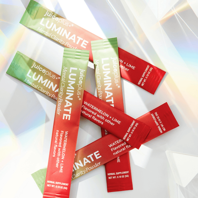 Several Juiceplus+ Luminate herbal clarity powder stick packs are arranged on a glossy white surface with soft rainbow reflections. The packets are shown in green, peach, and red, with white text reading LUMINATE and flavor notes including Watermelon + Lime.