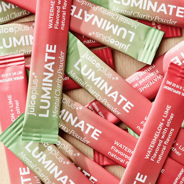 A close-up flat lay of Juiceplus+ LUMINATE Mental Clarity Powder sachets in red and green packaging, with white text, scattered on a light wood surface.