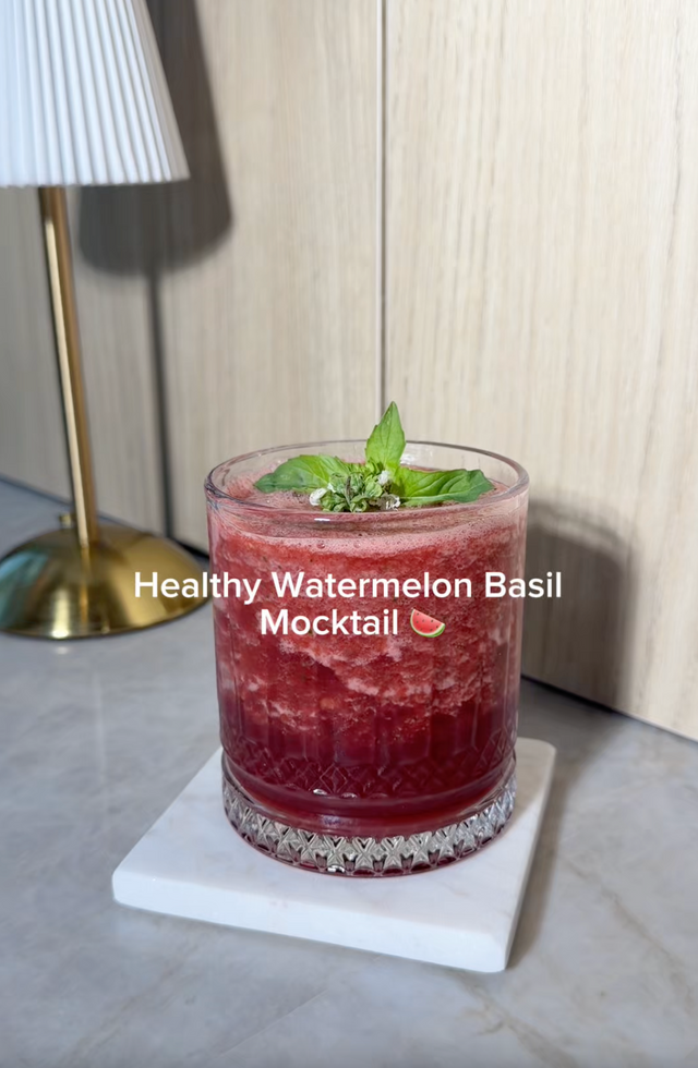 A glass of healthy watermelon basil mocktail cocktail on a white coaster, with a lamp and wood-paneled wall in the background. White text overlay reads 'Healthy Watermelon Basil Mocktail'.