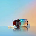 An amber glass supplement bottle lies on its side with two white capsules spilled in front of it on a reflective surface. The bottle label shows the brand name 'SUPER BLOOM' in white text against a blue-to-orange gradient background.