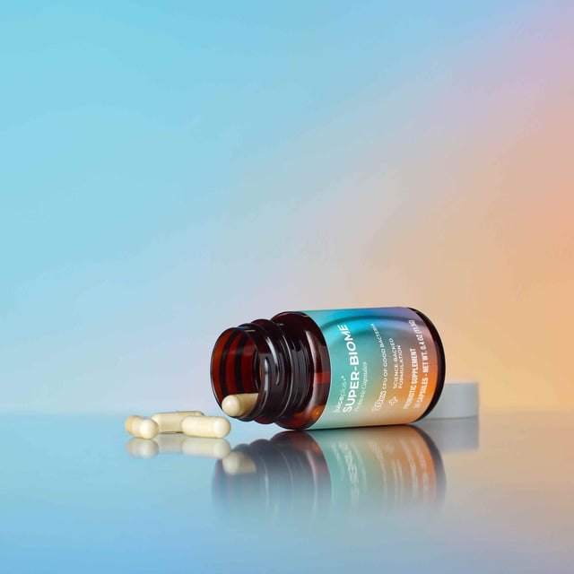 An amber glass supplement bottle lies on its side with two white capsules spilled in front of it on a reflective surface. The bottle label shows the brand name 'SUPER BLOOM' in white text against a blue-to-orange gradient background.