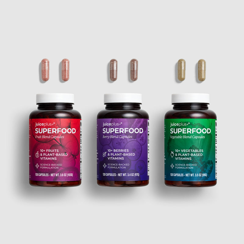 Three Juiceroop Superfood supplement bottles arranged side by side on a light gray background, each with three capsules floating above it. The bottles have white caps and dark brown bodies, with labels in red, purple, and green.